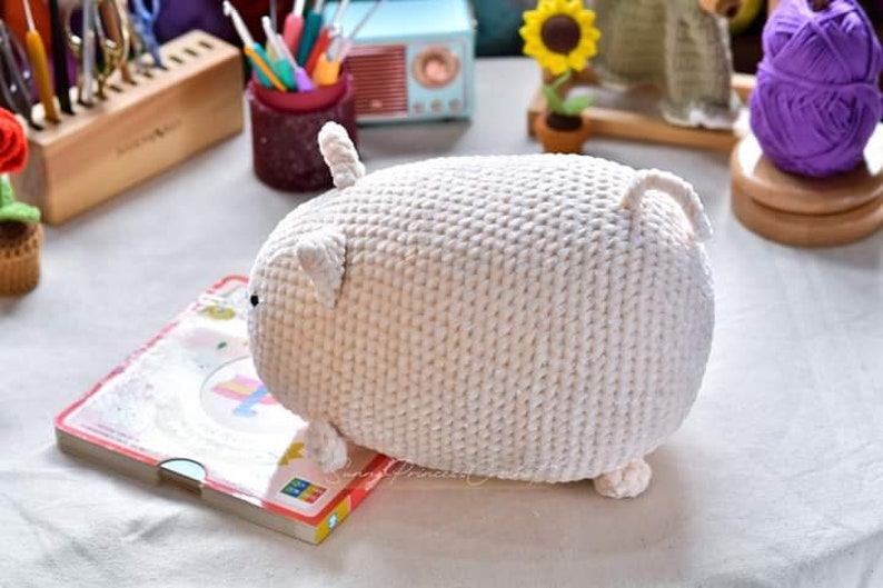 May include: White crocheted stuffed animal shaped like a pig. The pig has a small tail and two small ears.
