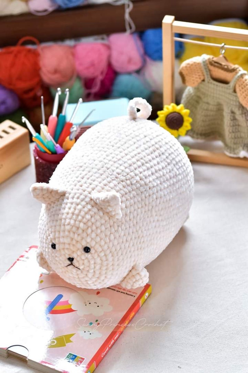 May include: White crocheted plush toy shaped like a pig with black eyes. The toy is sitting on a book with a rainbow and clouds design. The book has the text "Crafty Princess Crochet" on the cover.
