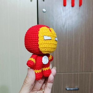 Puede incluir: Un juguete de peluche tejido a crochet de Iron Man. El juguete es rojo y amarillo con una cara blanca y un c&iacute;rculo blanco en el pecho. El juguete est&aacute; siendo sostenido en una mano.