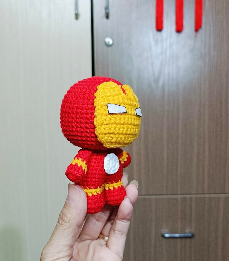 Puede incluir: Un juguete de peluche tejido a crochet de Iron Man. El juguete es rojo y amarillo con una cara blanca y un c&iacute;rculo blanco en el pecho. El juguete est&aacute; siendo sostenido en una mano.