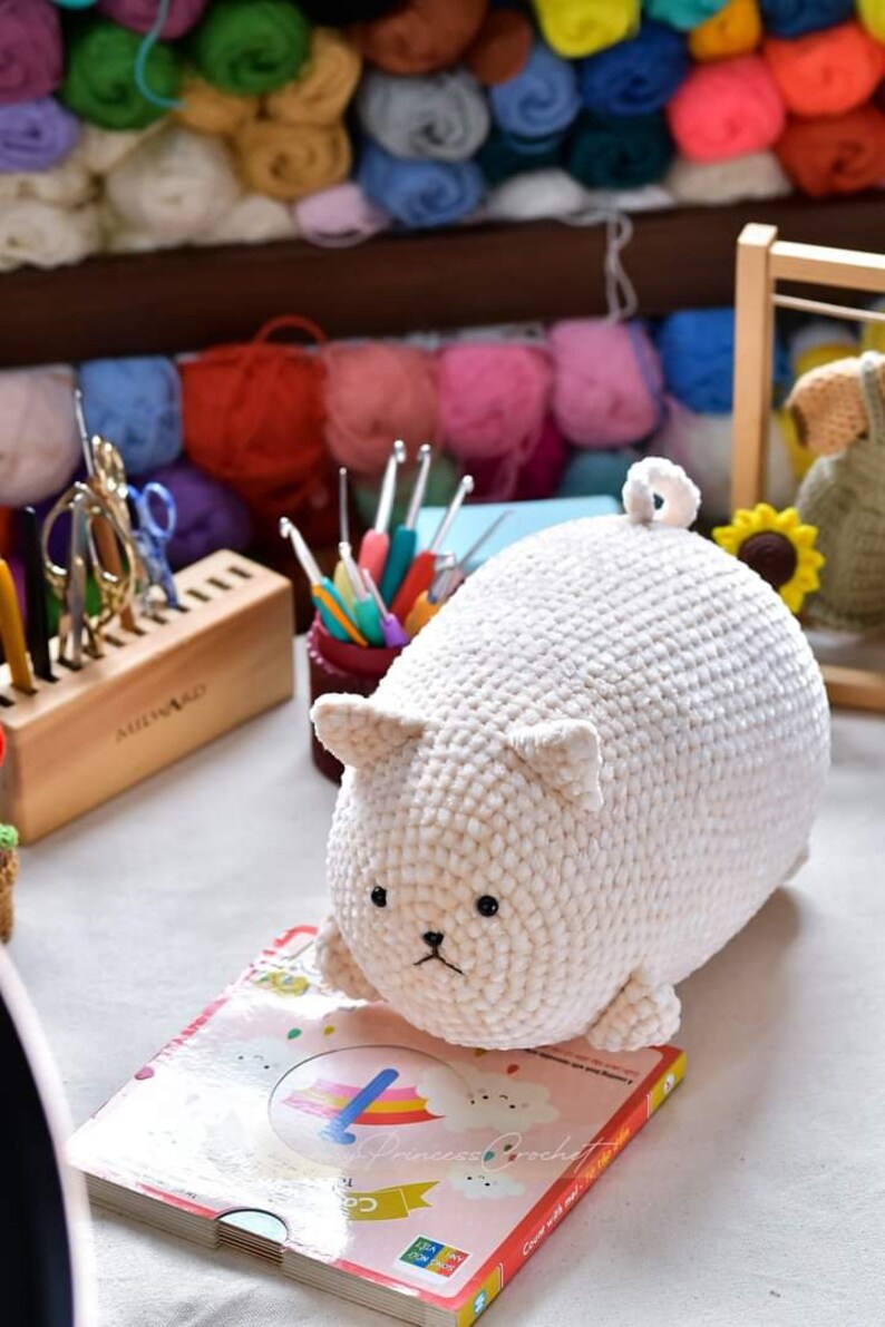 May include: A white crocheted plush toy shaped like a cat, with black eyes and a black nose. The toy is sitting on a book with a rainbow and clouds on the cover. The book says "Princess Crochet" and "Count with me, 1 to 10".