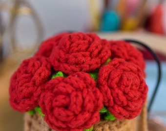 May include: A crocheted flower bouquet with red roses in a brown crocheted pot. The pot has a textured surface and the roses are arranged in a cluster.