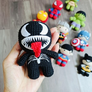 May include: A black crocheted Venom character with a red tongue and white eyes. The character is holding a web in its hand.