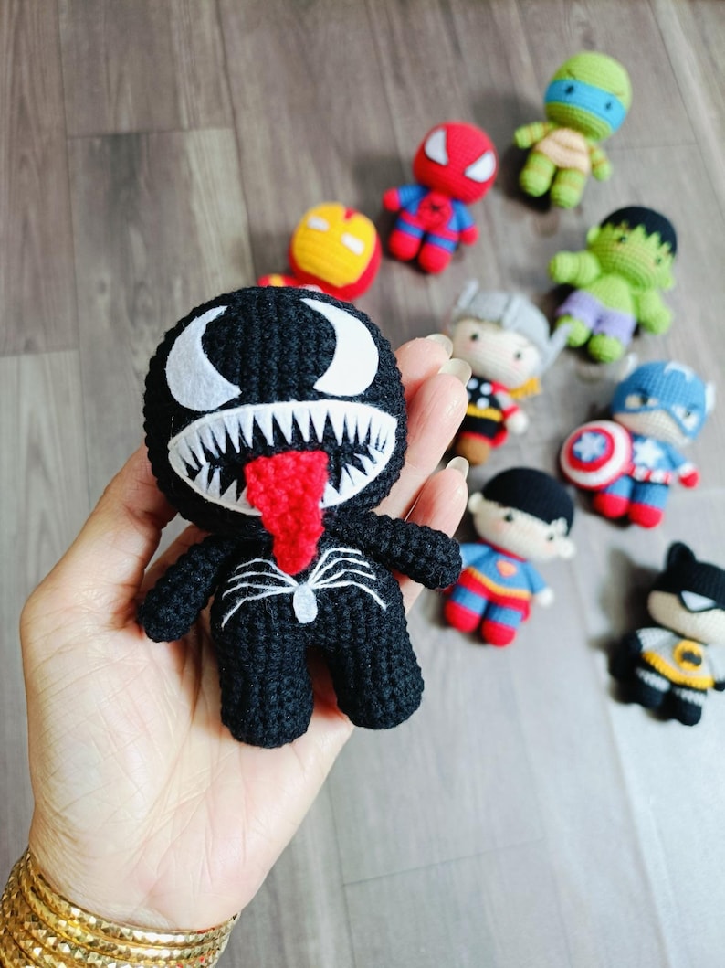 May include: A black crocheted Venom character with a red tongue and white eyes. The character is holding a web in its hand.