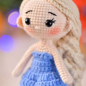 May include: A crocheted doll with long blonde hair wearing a blue dress. The doll has a sweet expression with big eyes and rosy cheeks.