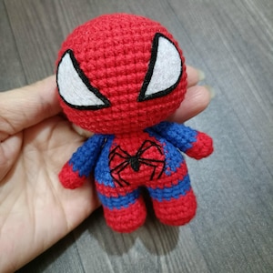 May include: A crocheted red and blue Spiderman toy with a black spider design on the chest. The toy has white eyes and a black outline.