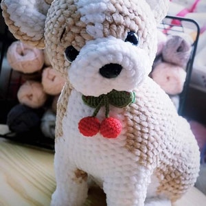 May include: A crocheted plush dog toy with a white and brown fur pattern. The dog is wearing a green crocheted collar with two red cherries hanging from it.