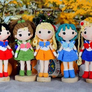 May include: Five crocheted dolls dressed as Sailor Moon characters. The dolls are standing on wooden bases. The dolls are wearing different colored outfits, including red, green, orange, blue, and light blue.