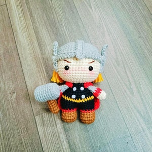 May include: A crocheted toy figure of Thor from the Marvel comics. The figure is wearing a grey helmet with yellow horns, a red and black costume, and holding a grey hammer.