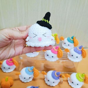 Puede incluir: Una colecci&oacute;n de adornos de fantasmas de crochet con sombreros de diferentes colores. Los fantasmas tienen caras sonrientes y sostienen peque&ntilde;as calabazas naranjas.