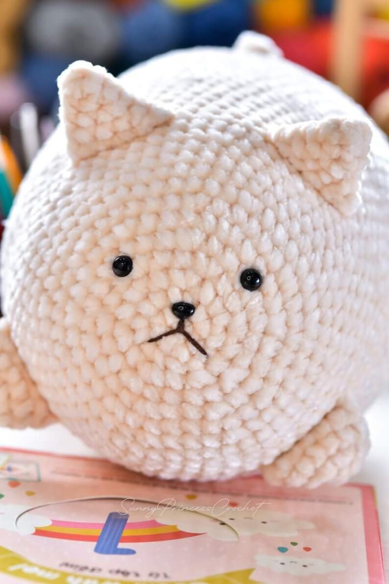 May include: A white crocheted plush cat toy with black eyes and a black nose. The cat is sitting on a colorful surface with the text "Sunny Princess Crochet".