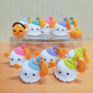 Puede incluir: Una colecci&oacute;n de llaveros de fantasmas de crochet con sombreros de diferentes colores y brazos naranjas. Los fantasmas tienen caras sonrientes y est&aacute;n dispuestos en un recipiente de pl&aacute;stico transparente. Hay una peque&ntilde;a calabaza de crochet en el recipiente.