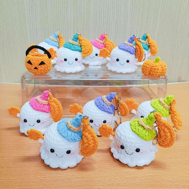 Puede incluir: Una colecci&oacute;n de llaveros de fantasmas de crochet con sombreros de diferentes colores y brazos naranjas. Los fantasmas tienen caras sonrientes y est&aacute;n dispuestos en un recipiente de pl&aacute;stico transparente. Hay una peque&ntilde;a calabaza de crochet en el recipiente.