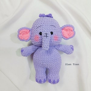 Puede incluir: Un elefante de peluche morado tejido a crochet con orejas rosas y un pequeño lazo en la cabeza. El elefante tiene ojos negros y una boca cosida. El juguete está sentado sobre una superficie blanca. @Lan Tran