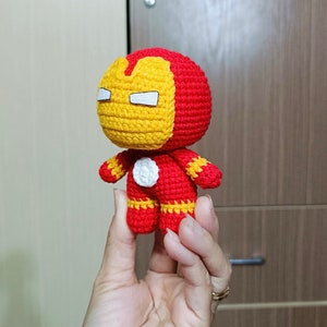 Puede incluir: Un juguete de peluche tejido a crochet de Iron Man. El juguete es rojo y amarillo con un c&iacute;rculo blanco en el pecho. El juguete est&aacute; siendo sostenido en la mano de una persona.
