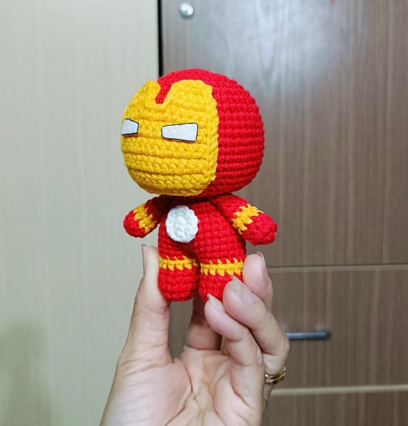 Puede incluir: Un juguete de peluche tejido a crochet de Iron Man. El juguete es rojo y amarillo con un c&iacute;rculo blanco en el pecho. El juguete est&aacute; siendo sostenido en la mano de una persona.
