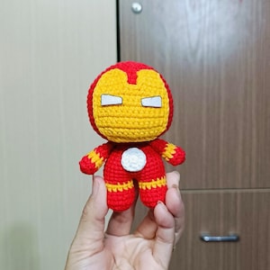 Puede incluir: Un juguete de peluche tejido a crochet de Iron Man, un superh&eacute;roe de Marvel Comics. El juguete es rojo y amarillo con un c&iacute;rculo blanco en el pecho. El juguete tiene las manos extendidas hacia adelante.