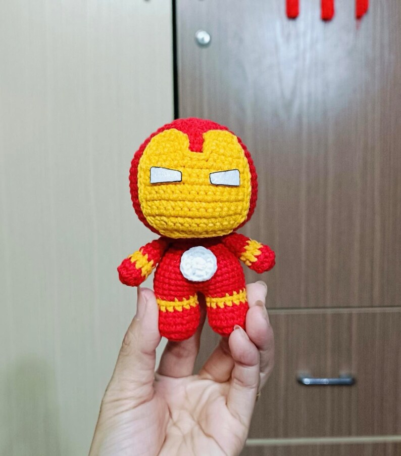 Puede incluir: Un juguete de peluche tejido a crochet de Iron Man, un superh&eacute;roe de Marvel Comics. El juguete es rojo y amarillo con un c&iacute;rculo blanco en el pecho. El juguete tiene las manos extendidas hacia adelante.