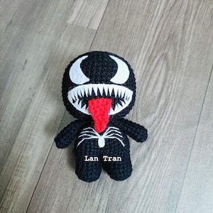 May include: Black and white crocheted Venom character with a red tongue. The character has a white spider symbol on its chest and the text "Lan Tran" is written below it.