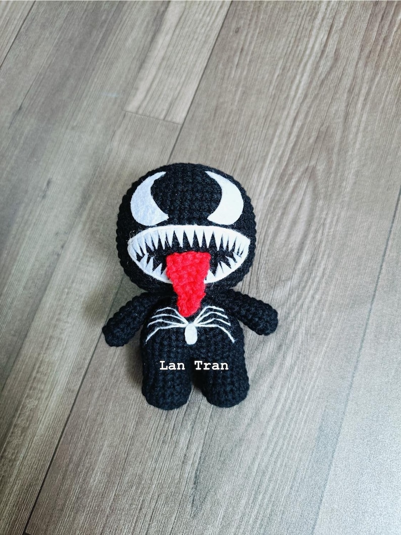 May include: Black and white crocheted Venom character with a red tongue. The character has a white spider symbol on its chest and the text "Lan Tran" is written below it.