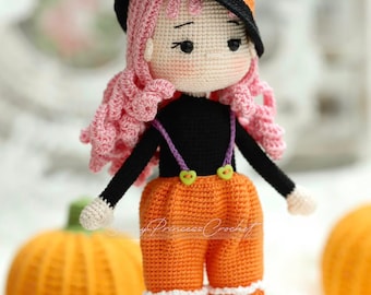 May include: A crocheted doll dressed as a witch for Halloween. The doll has pink hair, a black witch hat with an orange pumpkin on top, a black shirt, orange overalls, and purple shoes. The doll is standing in front of two orange pumpkins.