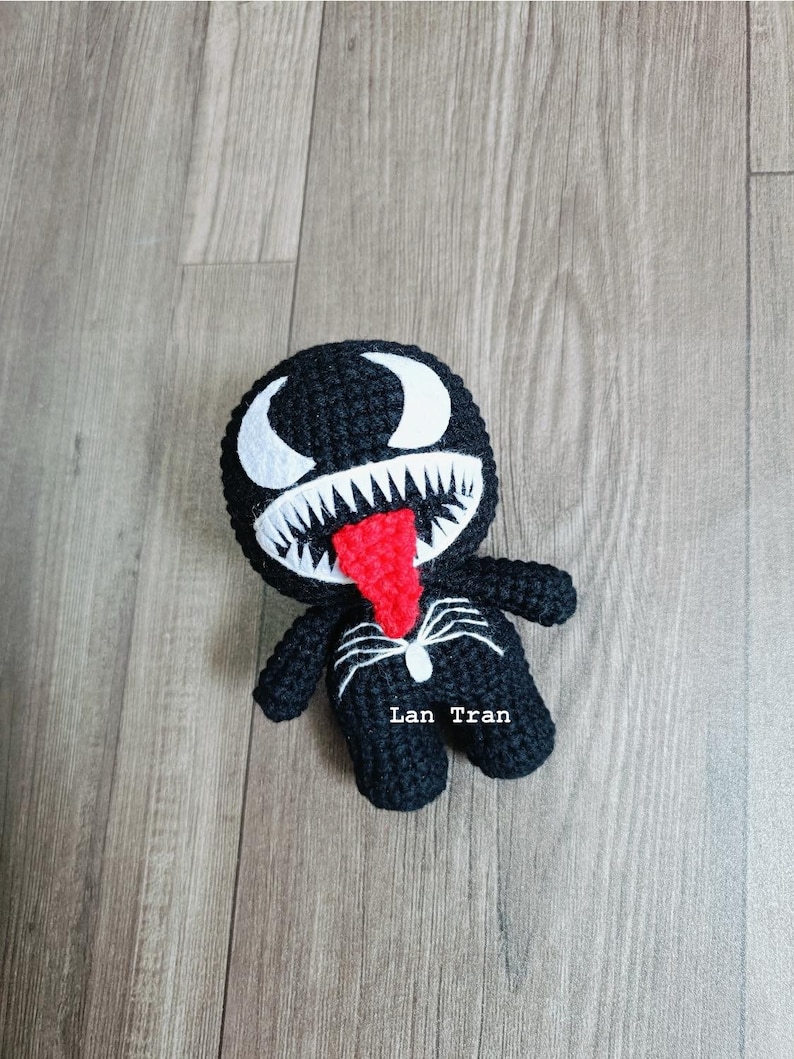 May include: Black crocheted Venom character with a red tongue and white eyes. The character is standing on a wood floor. The text "Lan Tran" is written on the character's chest.