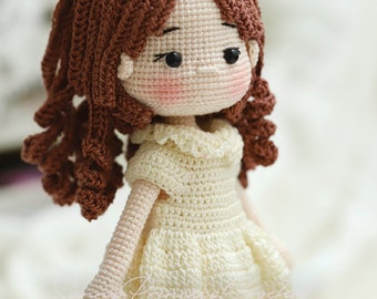 May include: A handmade crochet doll with brown curly hair and an orange bow. The doll wears a cream-colored dress with orange trim. The doll's face has rosy cheeks and black eyes. The doll is approximately 8 inches tall.