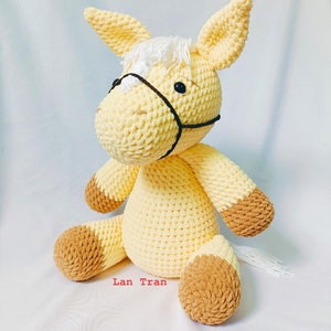 May include: A crocheted yellow horse toy with brown accents on the hooves and a white mane. The horse is sitting on a white surface.