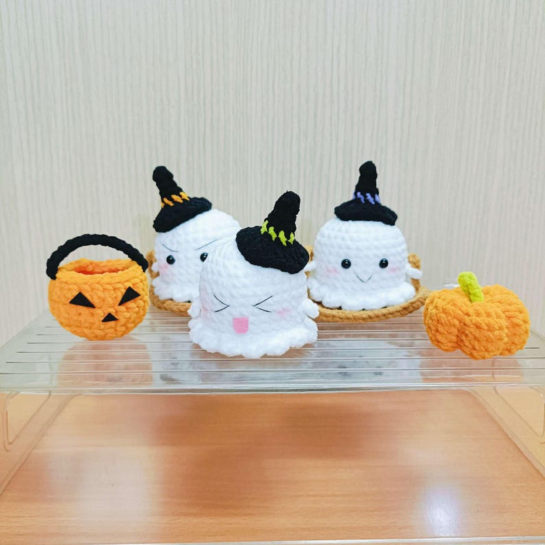 Puede incluir: Tres adornos de fantasmas de crochet con sombreros de bruja negros y una calabaza naranja de crochet. Los fantasmas tienen cuerpos de hilo blanco y ojos negros. La calabaza tiene un tallo verde.