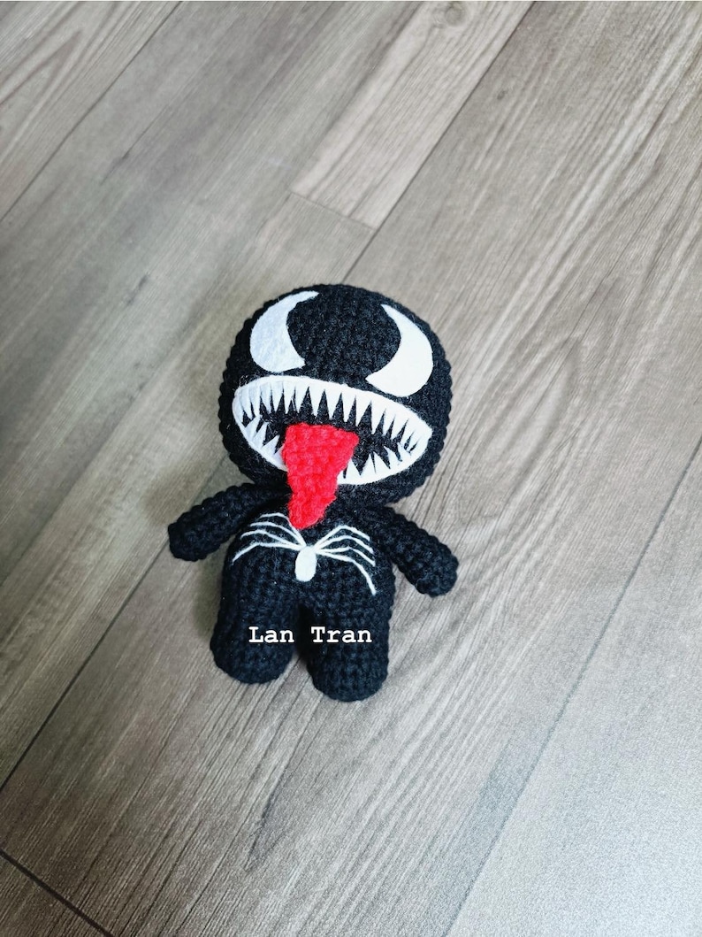 May include: Black crocheted Venom character plush toy with white eyes and teeth, a red tongue, and a spider web design on the chest. The toy is standing on a wood floor and has the text "Lan Tran" on the bottom.