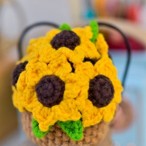 Könnte beinhalten: Ein gehäkelter Sonnenblumenkorb mit leuchtend gelben Blütenblättern, dunkelbraunen Zentren und grünen Blättern. Der Korb ist hellbraun und hat einen schwarzen Griff. Der Text "Sunny Princess Crochet" ist auf dem Korb sichtbar.