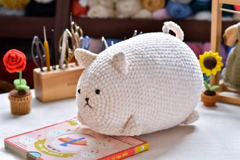 May include: A white crocheted stuffed animal shaped like a cat. The cat has a round body and small black eyes. The cat is sitting on a book with a rainbow design.