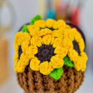 Puede incluir: Una maceta de girasol de ganchillo hecha con hilo marr&oacute;n, amarillo y verde. La maceta est&aacute; sobre una superficie de madera.