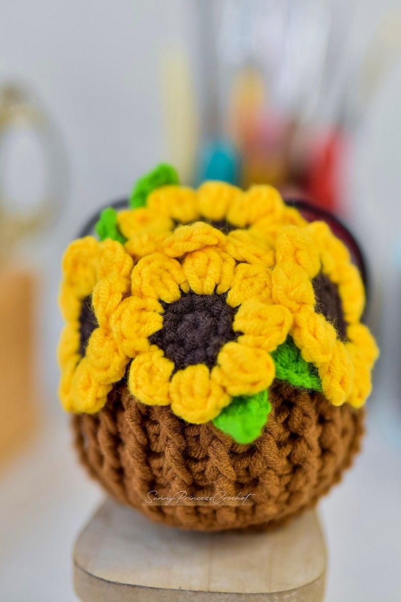 Puede incluir: Una maceta de girasol de ganchillo hecha con hilo marr&oacute;n, amarillo y verde. La maceta est&aacute; sobre una superficie de madera.