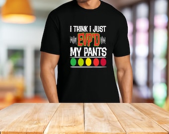 May include: Black t-shirt with the text "I THINK I JUST EVP'D MY PANTS" in white and red. Below the text are four colored circles: green, yellow, and red. The shirt is worn by a person.