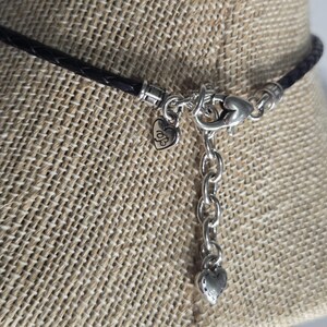 以下が含まれることがあります： 黒い編み込みレザーネックレスのクローズアップ。シルバーのハート型チャームとチェーンエクステンダーが付いています。 ネックレスはロブスタークラスプ留めで、チェーンの端には小さなハートチャームが付いています。