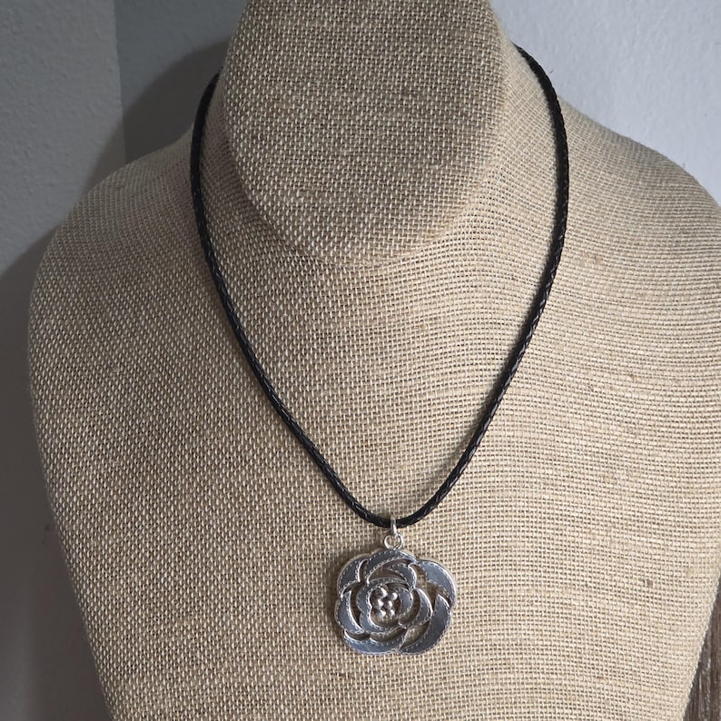 以下が含まれることがあります： 黒いコードに銀色のバラのペンダントネックレス。ペンダントは、バラの繊細な透かし彫りデザインです。ネックレスは、ベージュの布で覆われたマネキンに展示されています。