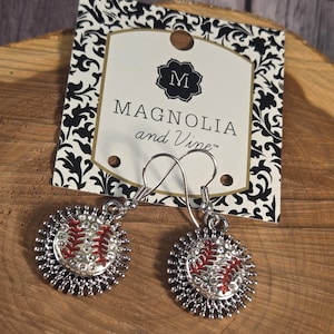 Puede incluir: Pendientes de béisbol plateados con costuras rojas y detalles de cristal transparente. Los pendientes están sobre una superficie de madera con una etiqueta de Magnolia and Vine.