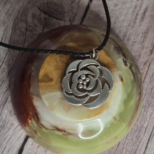 以下が含まれることがあります： バラのデザインが施されたシルバーカラーのペンダントネックレス。ペンダントは黒い編み込みコードから吊り下げられています。ネックレスは、緑と茶色の石の表面に、木製の背景で展示されています。