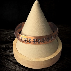 May include: A copper cuff bracelet with a geometric pattern of triangles and circles. The bracelet is displayed on a beige cone-shaped stand, set against a dark background. The bracelet's design features cut-out shapes.