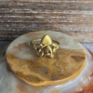Puede incluir: Un anillo dorado con un diseño de pulpo. El anillo tiene una figura detallada de pulpo con tentáculos que se envuelven alrededor de la banda y una cabeza en la parte superior. El anillo está sobre una superficie de piedra con un fondo de madera.