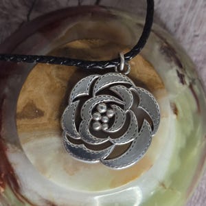以下が含まれることがあります： バラのデザインが施されたシルバーカラーのペンダントネックレス。ペンダントは黒色の編み込みコードから吊り下げられています。バラはステッチのような外観で、中央に小さく丸いアクセントがあります。ジュエリーです。