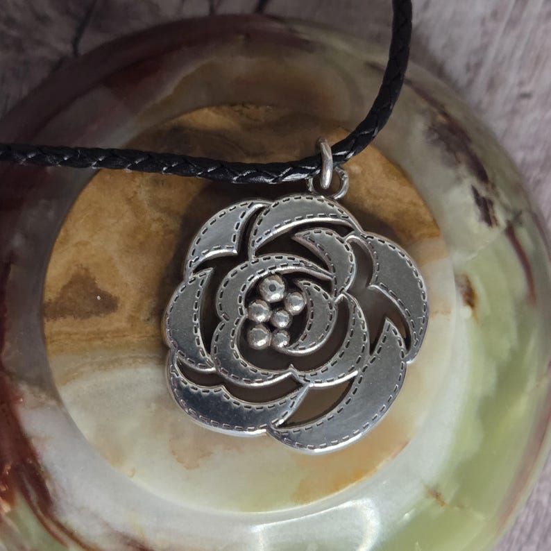 以下が含まれることがあります： バラのデザインが施されたシルバーカラーのペンダントネックレス。ペンダントは黒色の編み込みコードから吊り下げられています。バラはステッチのような外観で、中央に小さく丸いアクセントがあります。ジュエリーです。