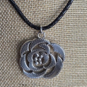 以下が含まれることがあります： 詳細な花の模様が施されたシルバーカラーのペンダントネックレス。ペンダントは黒色の編み込みコードから吊り下げられています。花のペンダントは、質感のある表面と、小さな丸いディテールの中心的なクラスターを備えています。