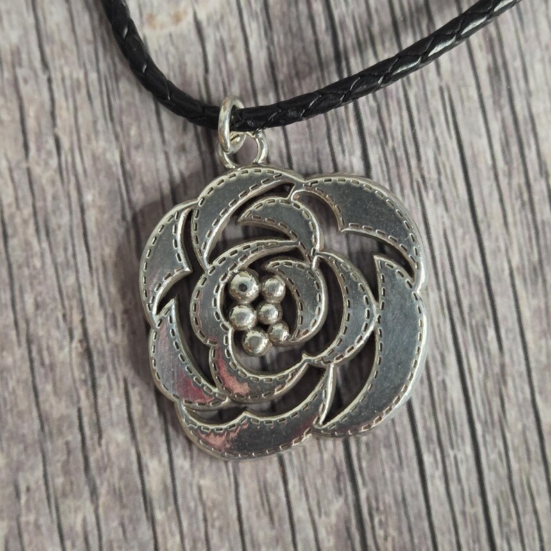 以下が含まれることがあります： 詳細なバラのデザインが施されたシルバーカラーのペンダントネックレス。ペンダントは黒色の編み革紐から吊り下げられています。バラはステッチの縁取りと中心部の小さな球体のクラスターを備えたテクスチャード加工の外観をしています。
