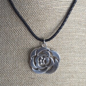 以下が含まれることがあります： 黒いコードに銀色のバラのペンダントネックレス。ペンダントは、テクスチャ加工されたエッジと小さな円の中心クラスターを備えた詳細な花のデザインが特徴です。ネックレスは、ニュートラルなテクスチャ加工された背景に対して表示されます。