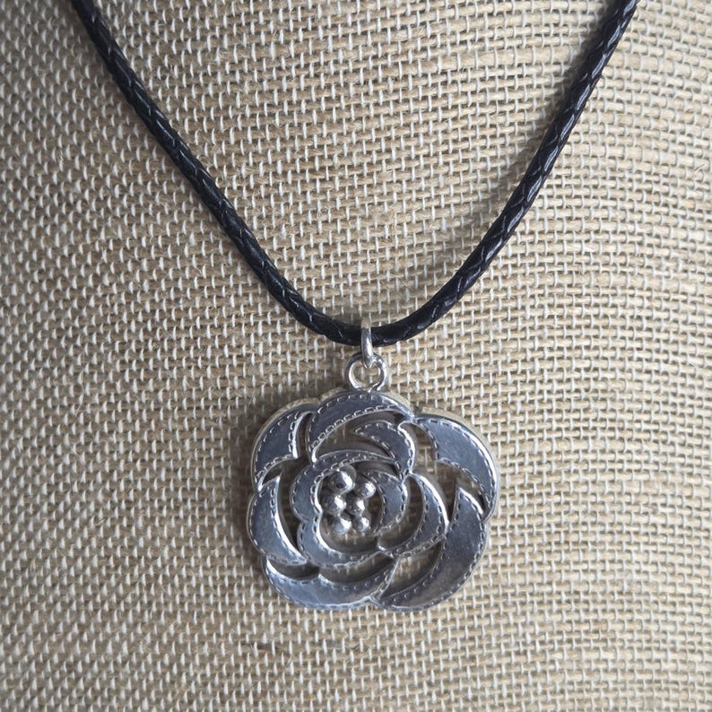 以下が含まれることがあります： 黒いコードに銀色のバラのペンダントネックレス。ペンダントは、テクスチャ加工されたエッジと小さな円の中心クラスターを備えた詳細な花のデザインが特徴です。ネックレスは、ニュートラルなテクスチャ加工された背景に対して表示されます。