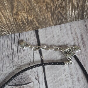 以下が含まれることがあります： 黒い編み革紐が付いた、シルバー色のネックレスの留め金とチェーンのクローズアップ。留め金にはハート型のアクセントが付いています。背景は明るい色の木製の表面です。