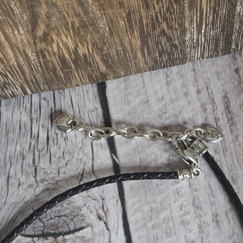 以下が含まれることがあります： 黒い編み革紐が付いた、シルバー色のネックレスの留め金とチェーンのクローズアップ。留め金にはハート型のアクセントが付いています。背景は明るい色の木製の表面です。