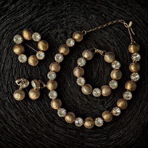 Könnte beinhalten: Ein Vintage-Schmuckset mit einer goldfarbenen Halskette, einem Armband, Ohrringen und einer Brosche. Der Schmuck hat strukturierte goldene Perlen, die mit klaren, funkelnden Strasssteinen durchsetzt sind. Das Set ist auf einer dunklen, strukturierten Oberfläche angeordnet.